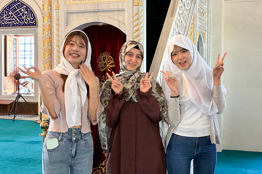 東京ジャーミイの礼拝堂で、白いスカーフを着けた敬愛大学国際学部1年庄司ゼミの女子学生2人と女性が並ぶ。首都圏でイスラム、トルコ、ネパールの文化を五感で学んだゼミ旅行の一場面。