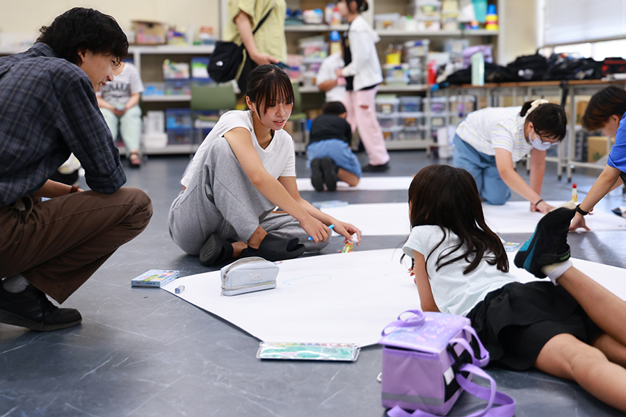 図工ワークショップで、大きな紙を前に小学生と目線を合わせ、描きたい巨大生命体のイメージを指さしながら確認する敬愛大学教育学部久保田ゼミの学生。周囲でも学生が小学生一人ひとりの制作を支援している。