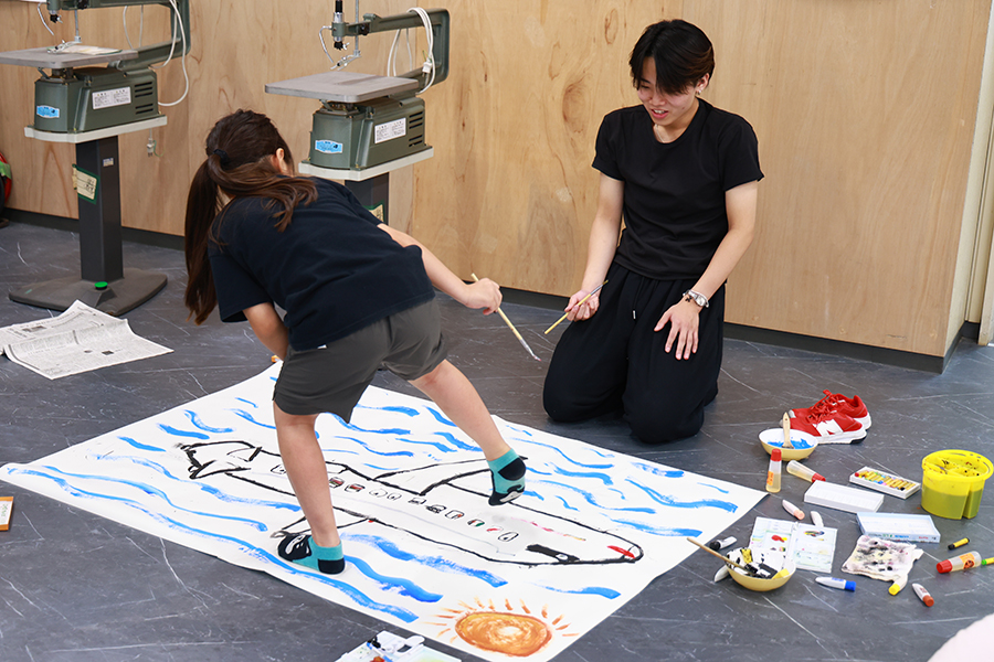 魚のような巨大生命体に青い線を描き加える小学生と、膝をついて目線を合わせ、筆を持ちながら相談する敬愛大学教育学部久保田ゼミの学生。対話と見守りを通じて制作を支えている。