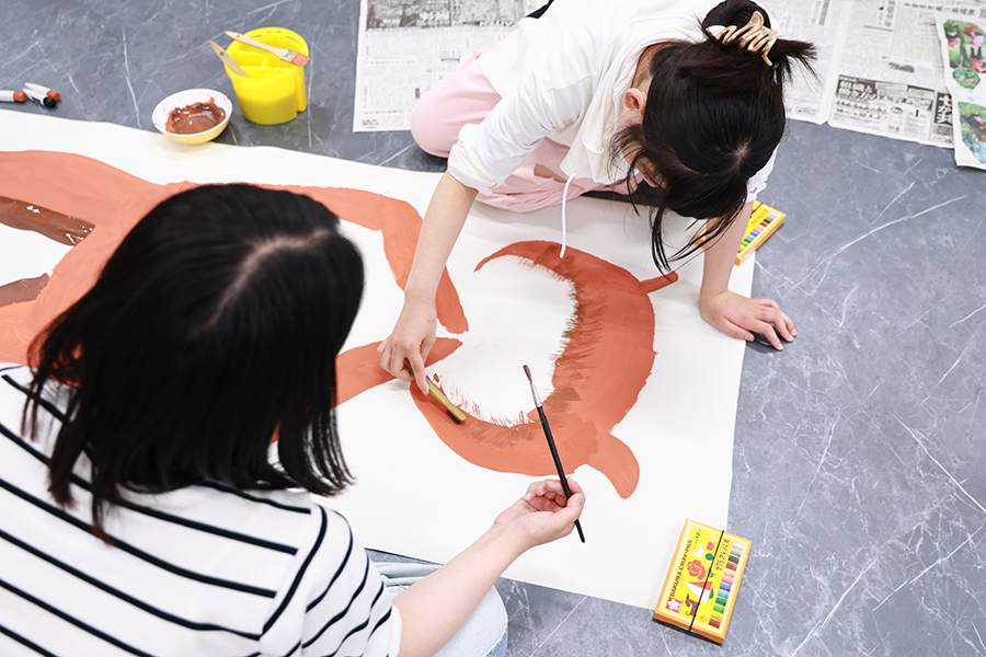 大きな紙に猫のような巨大生命体を描く小学生が、歯ブラシに茶色の絵の具を付けて毛並みを表現し、敬愛大学教育学部久保田ゼミの学生が筆を持って制作を見守っている。